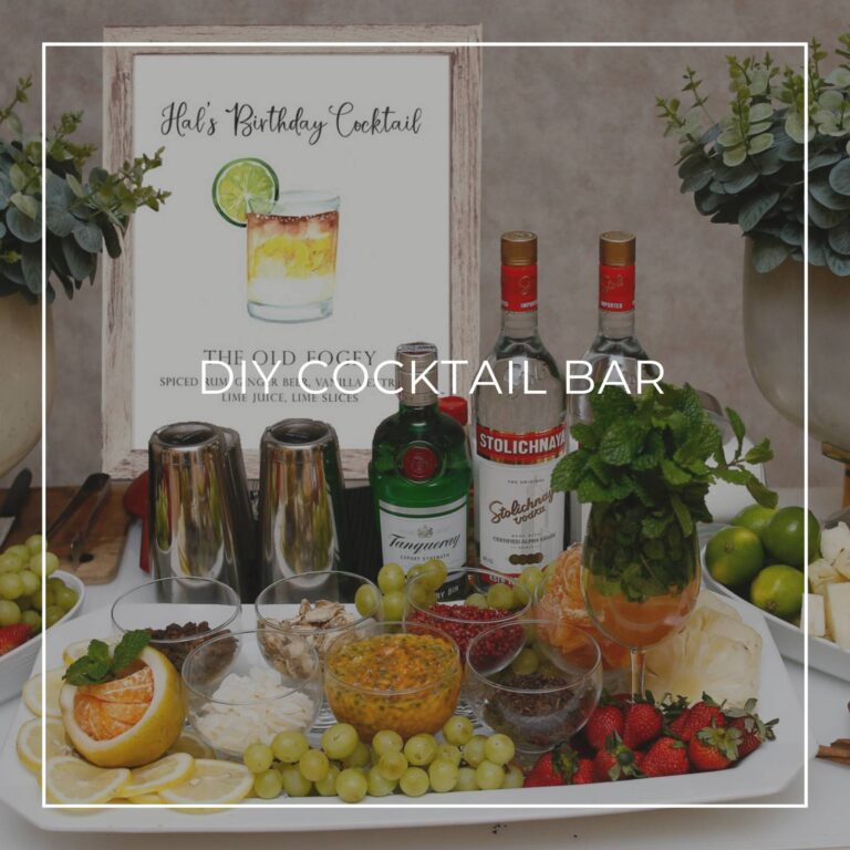 DIY cocktail bar with liquor, ingredients, and a cocktail recipe sign for party guests to make their own drink