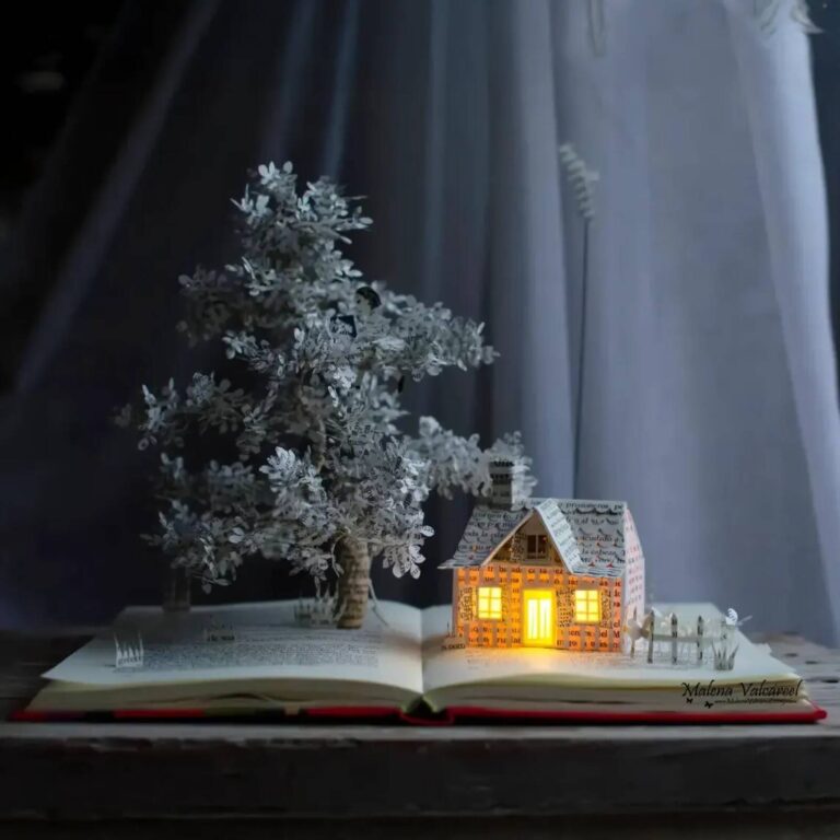Glowing paper cottage sculpture beside a leafless tree with a picket fence and paper birds, on an open book