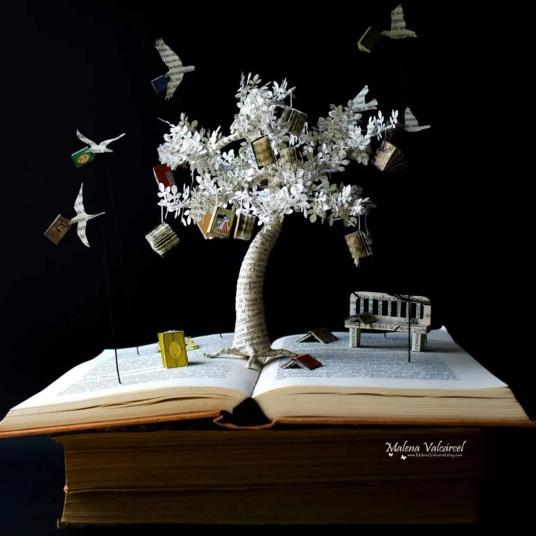 Book art sculpture of a tree hung with tiny miniature books and paper birds, with a bench, on an open book