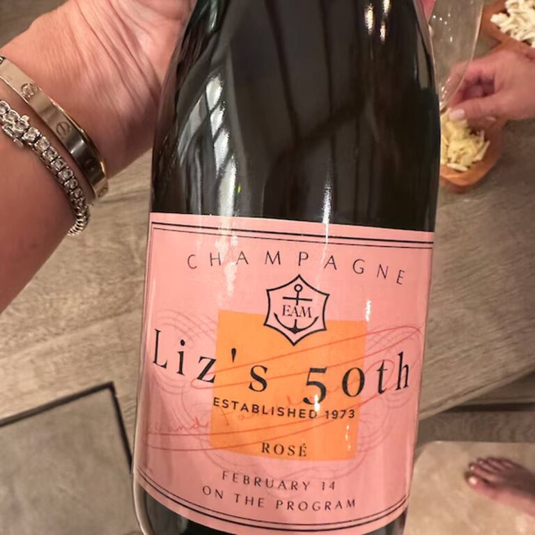 Hand holding a champagne bottle with a pink "Liz's 50th, Established 1973, Rosé" label, cheese board in the background