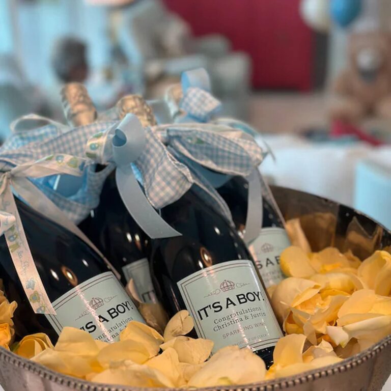 Champagne bottles wrapped in blue gingham ribbon with "It's a Boy! Congratulations Christina & Justin" labels, nestled in yellow rose petals