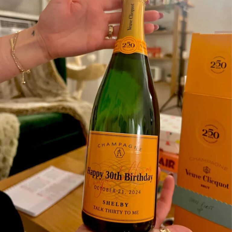 Hand holding a bottle with an orange "Happy 30th Birthday, Shelby, Talk Thirty to Me" label, a Veuve Clicquot 250th anniversary gift box behind it