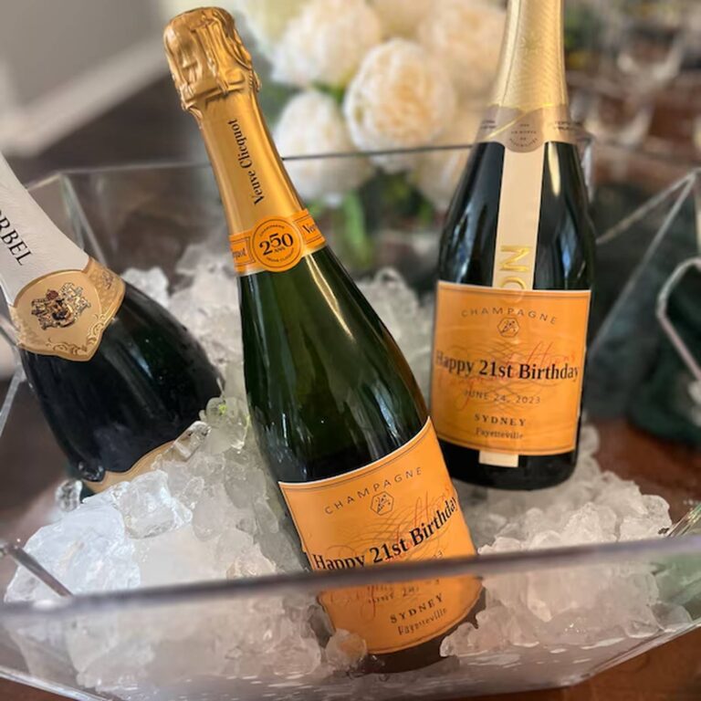 Two champagne bottles in an ice bucket with orange "Happy 21st Birthday, Sydney" labels, white roses in the background