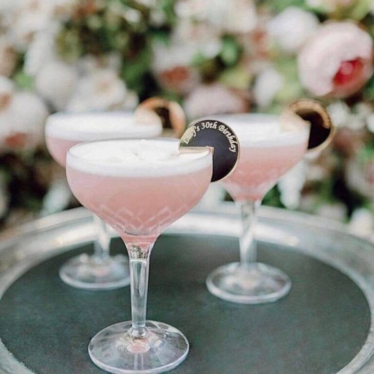 Two pink cocktails in coupe glasses with round black-and-gold drink charms reading “Lucy’s 30th Birthday”