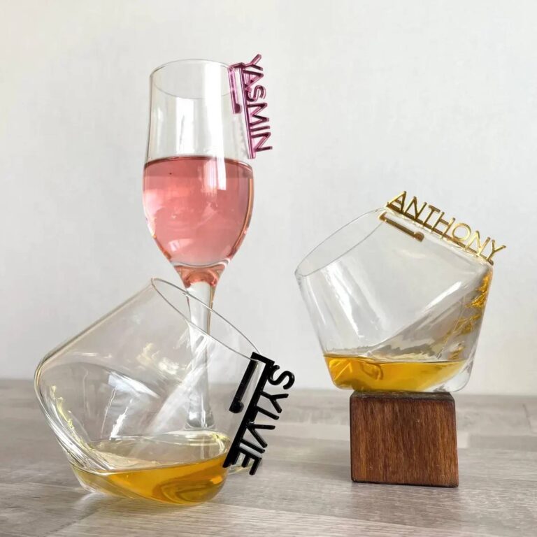 Two tumbler glasses with acrylic name charms reading “Yasmin” in pink and “Anthony” in gold, a wine glass with a “Sylvie” charm in front