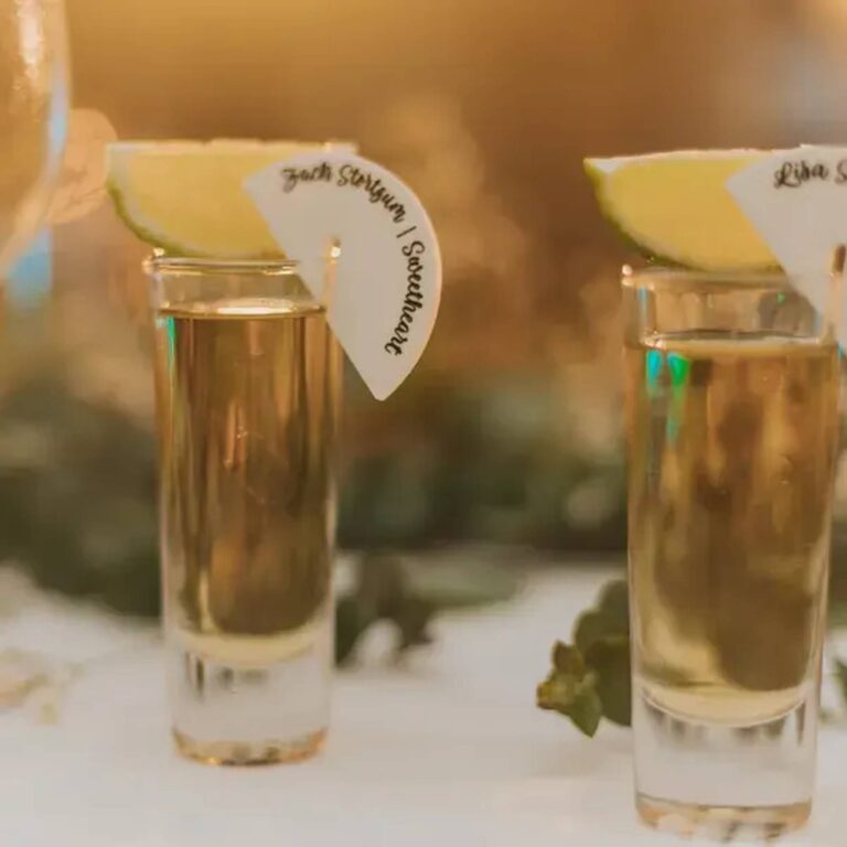 Two tequila shots with lime garnish and fan-shaped paper drink charms naming wedding party members, including “Zach Sturgeon, Sweetheart”
