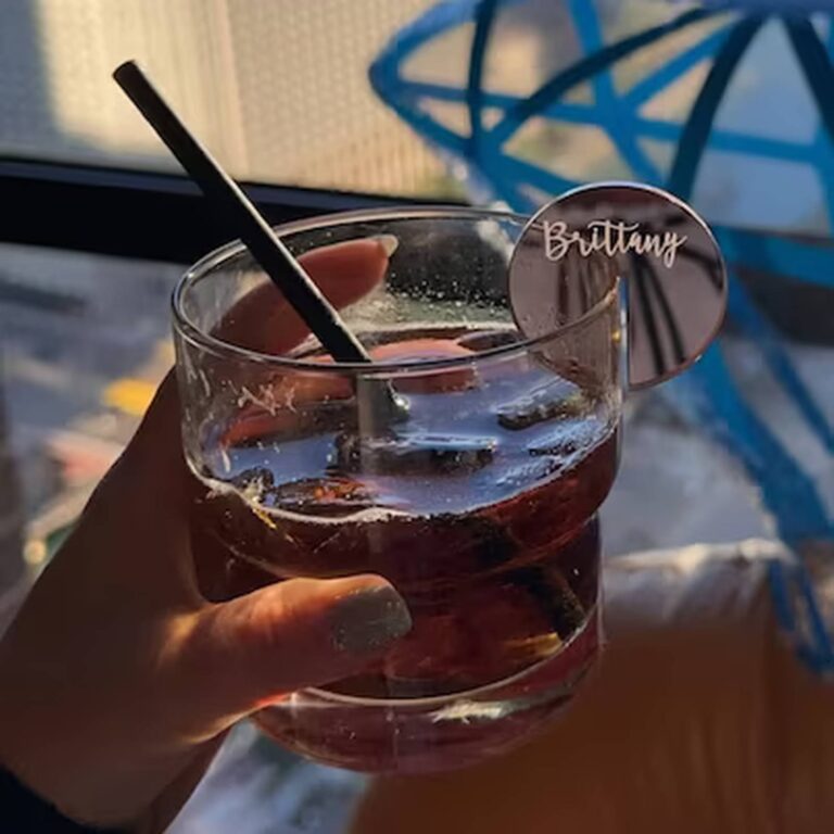 Hand holding a dark cocktail with a black straw and a round mirrored drink charm engraved “Brittany”