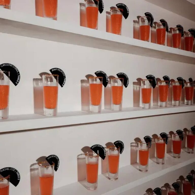 Wall of shot glasses filled with orange cocktails, each tagged with a black fan-shaped charm printed with a guest’s name and table number