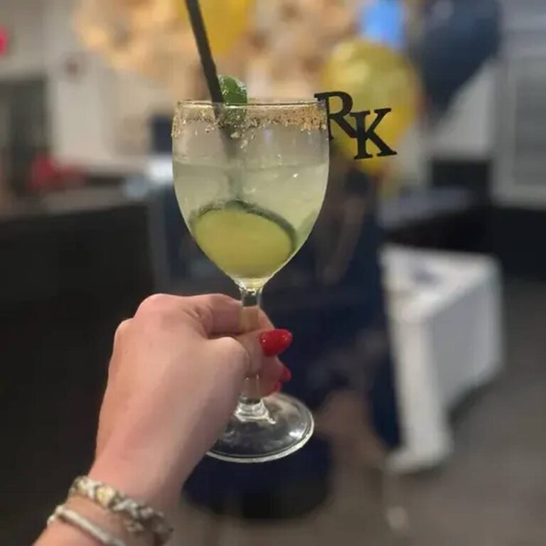 Hand holding a margarita with a gold salt rim and a black acrylic “RK” monogram drink charm, balloons in the background