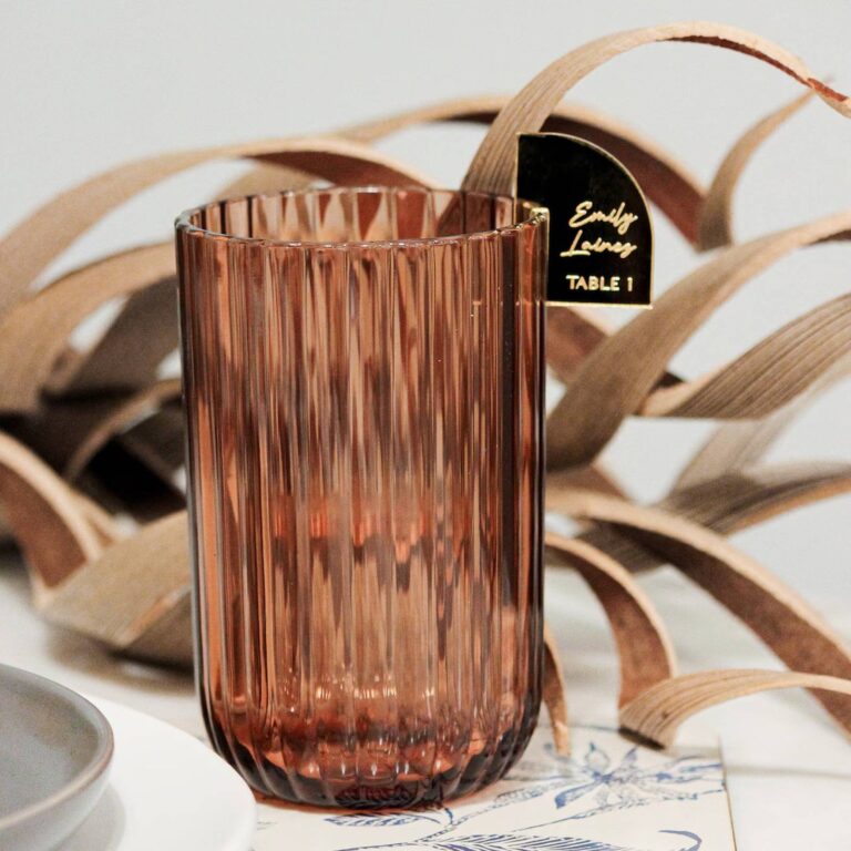 Ribbed amber glass with a gold arc-shaped drink charm reading “Emily Laines, Table 1,” woven decor in the background