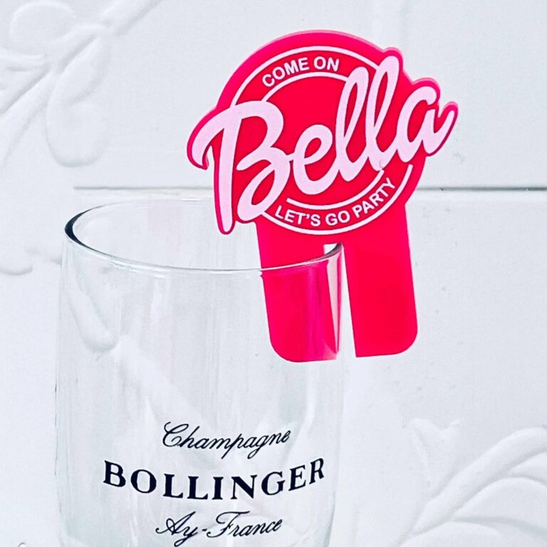 Close-up of a Bollinger champagne glass with a hot pink drink charm reading “Come On Bella, Let’s Go Party”