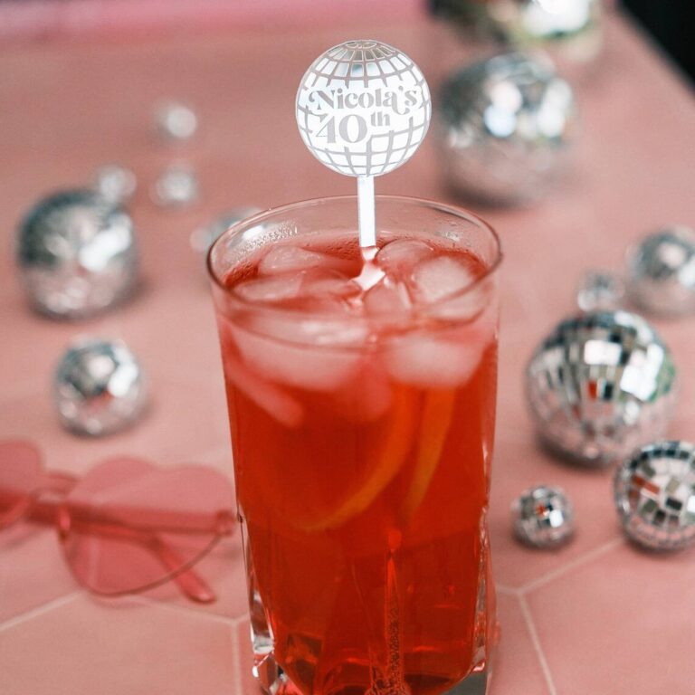 Red cocktail with a disco-ball-shaped acrylic drink stirrer engraved “Nicola’s 40th,” styled with disco balls and pink sunglasses