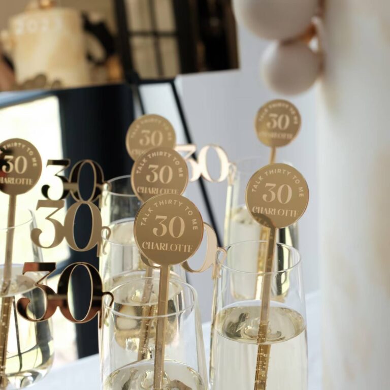 Champagne flutes with gold mirrored acrylic drink stirrers reading “Talk Thirty to Me, 30, Charlotte”