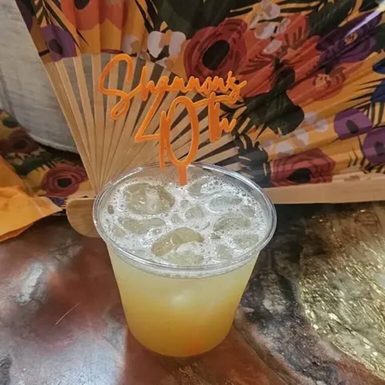 Iced drink in a plastic cup with an orange acrylic stirrer reading “Shannon’s 40th” against a floral fan backdrop