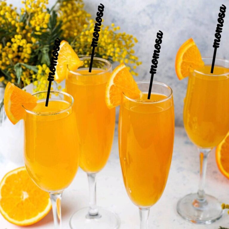 Champagne flutes of mimosa with black acrylic stirrers reading “mimosa,” garnished with orange wedges