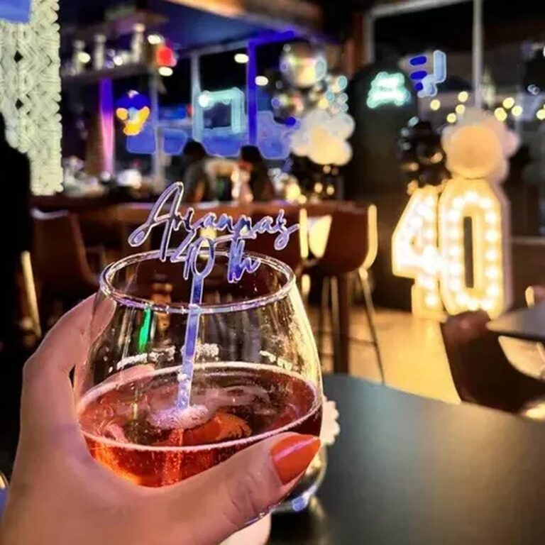 Wine glass held in hand with a clear acrylic stirrer reading “Arianna’s 40th” in a bar lit with neon signs and a 40 marquee