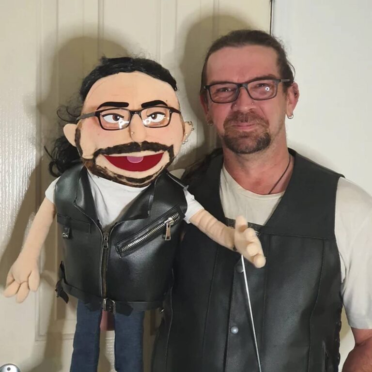 Man with long dark hair and goatee poses in a leather vest next to his matching biker-style custom puppet