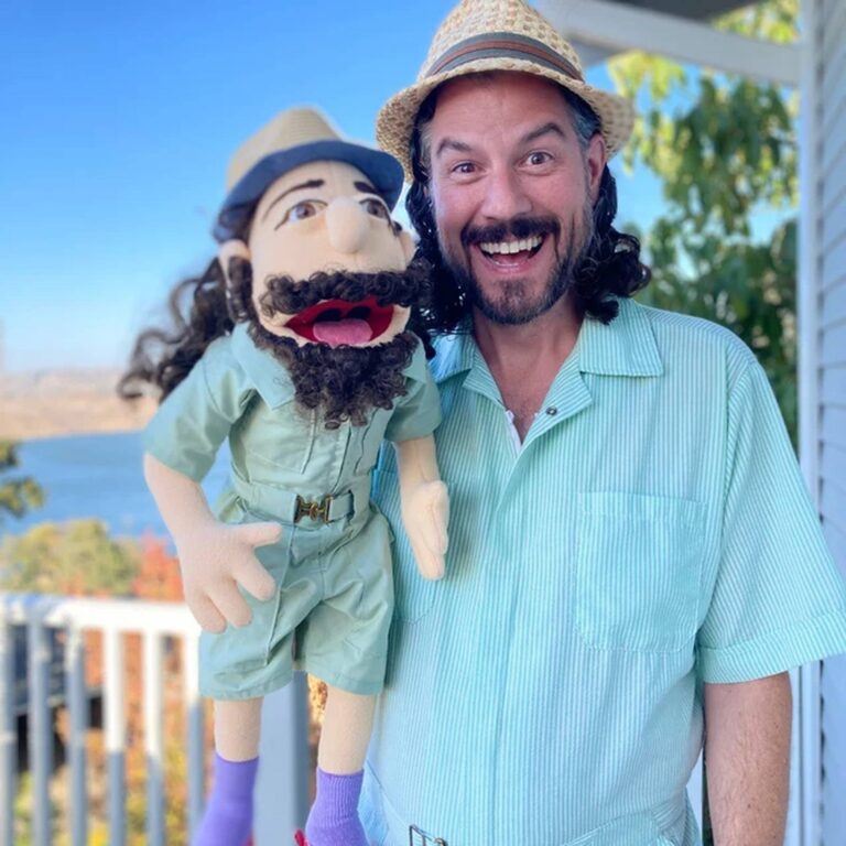 Man in a straw hat and teal shirt holds his safari-outfit puppet twin on a lakeside porch