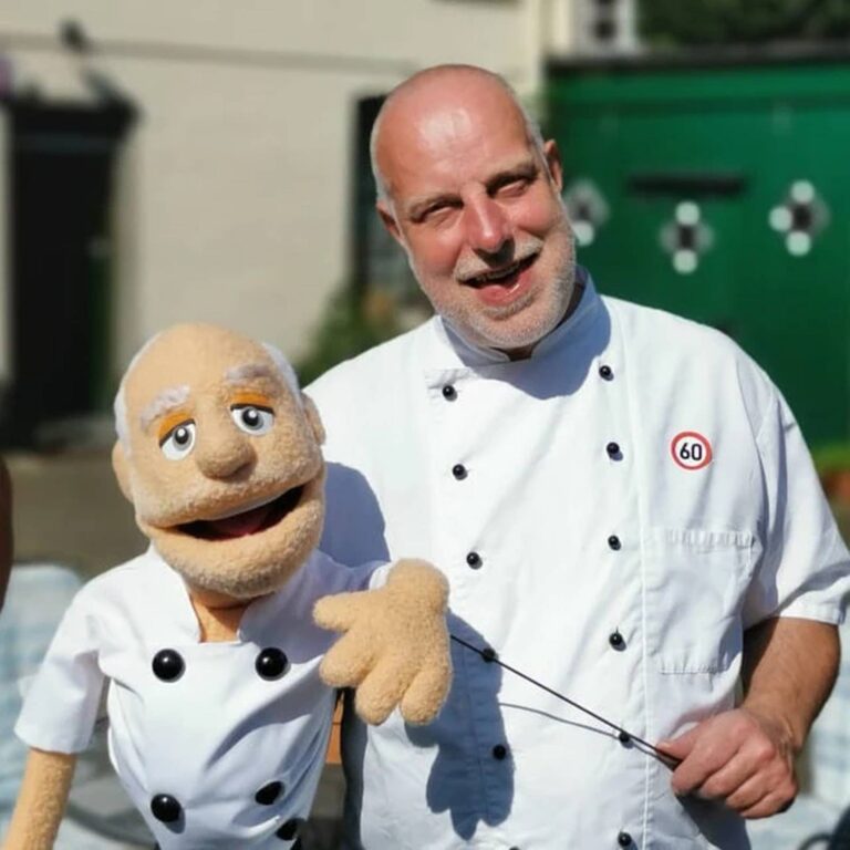Bald chef in a white chef’s coat holds his matching bald chef puppet outdoors near a green shed