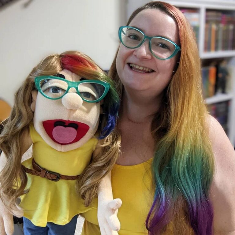 Woman with rainbow-dyed hair and teal glasses holds her matching puppet twin in a yellow top