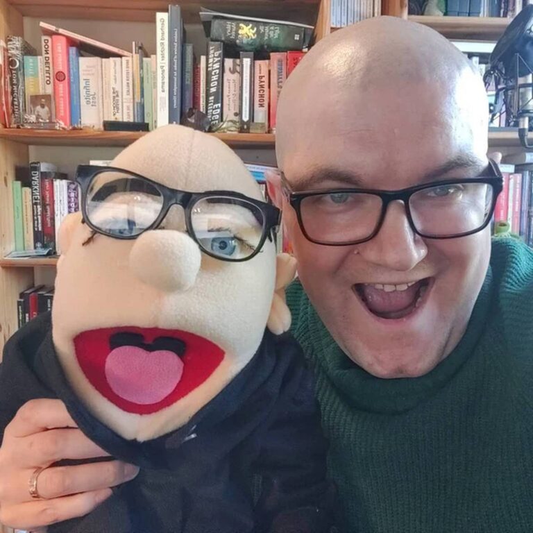 Bald man in a green sweater and black glasses smiles beside his matching bald puppet
