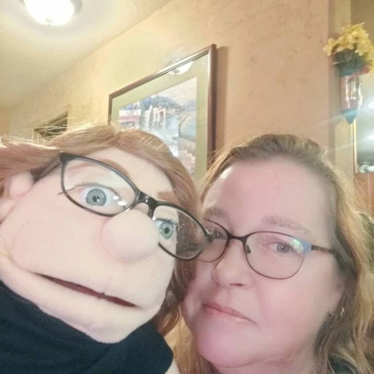 Two women share a selfie, one holding an auburn-haired puppet wearing glasses