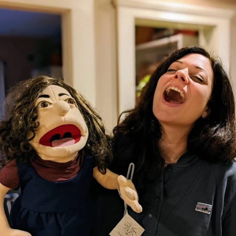 Laughing woman with dark curly hair holds her matching puppet twin in a navy dress