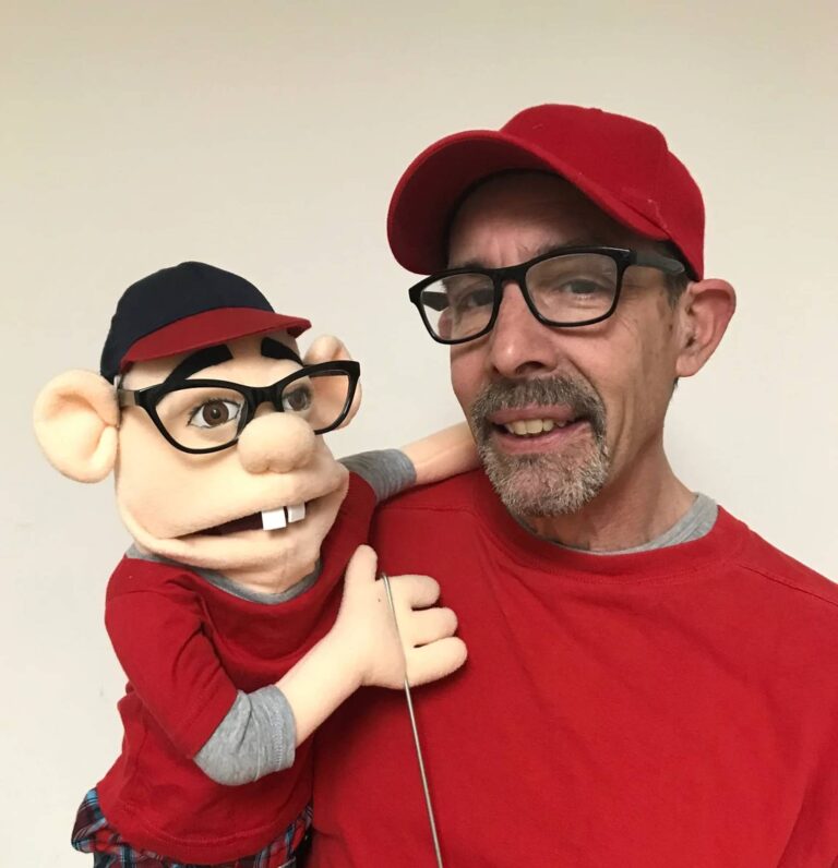Man in a red cap and shirt holds his gap-toothed puppet twin, also dressed in red