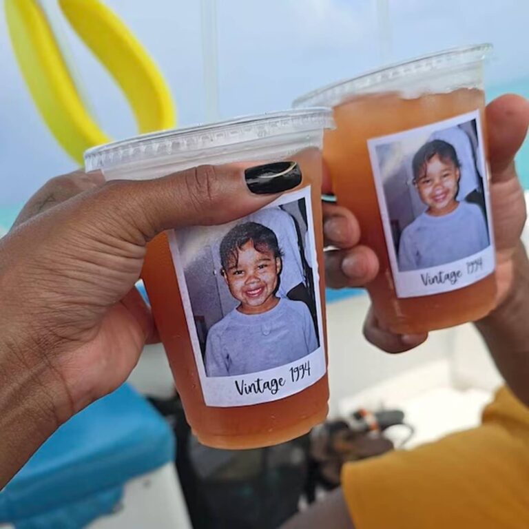 Two hands toasting with orange drinks in photo cups printed with a smiling toddler’s photo and “Vintage 1994,” beach and pool float in the background