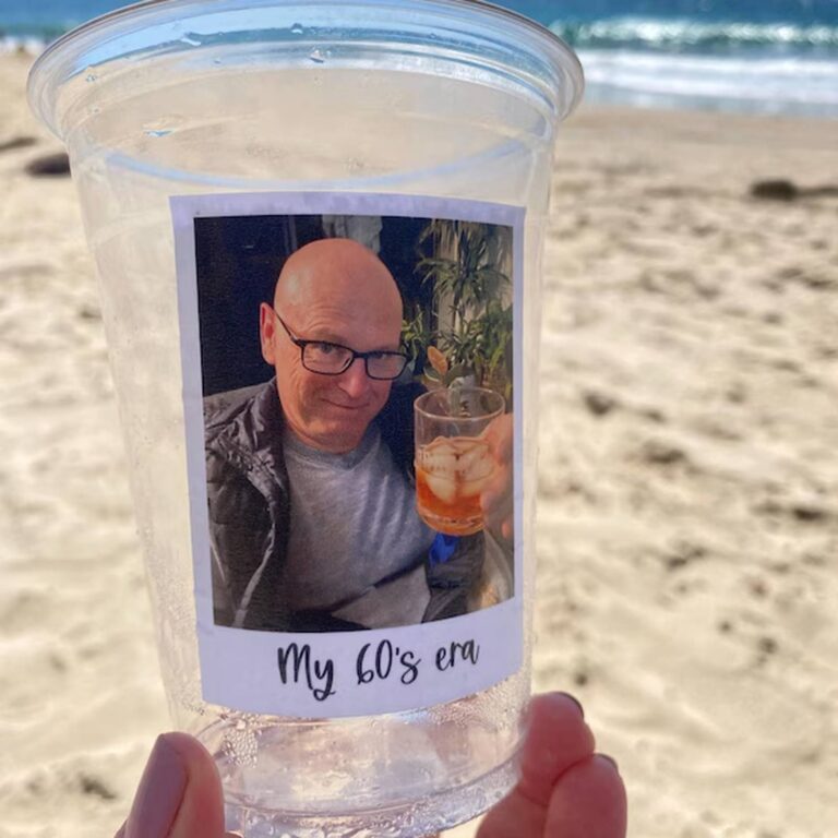Custom photo cup held up at the beach with a picture of a bald man in glasses holding a cocktail, captioned “My 60’s era”
