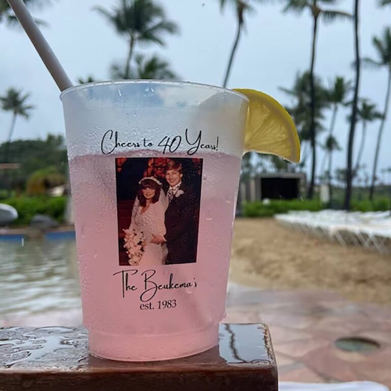 Pink cocktail with a lemon wedge in a custom photo cup reading “Cheers to 40 Years! The Beukema’s est. 1983” with a wedding photo, poolside with palm trees