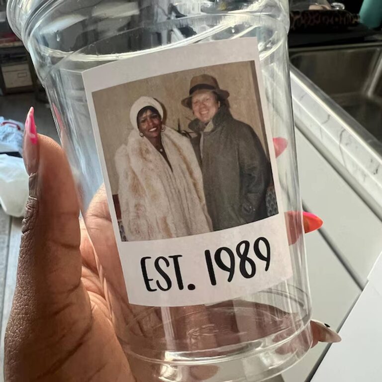 Photo cup held in a hand with pink nails, printed with a photo of two women in fur coats and “Est. 1989”