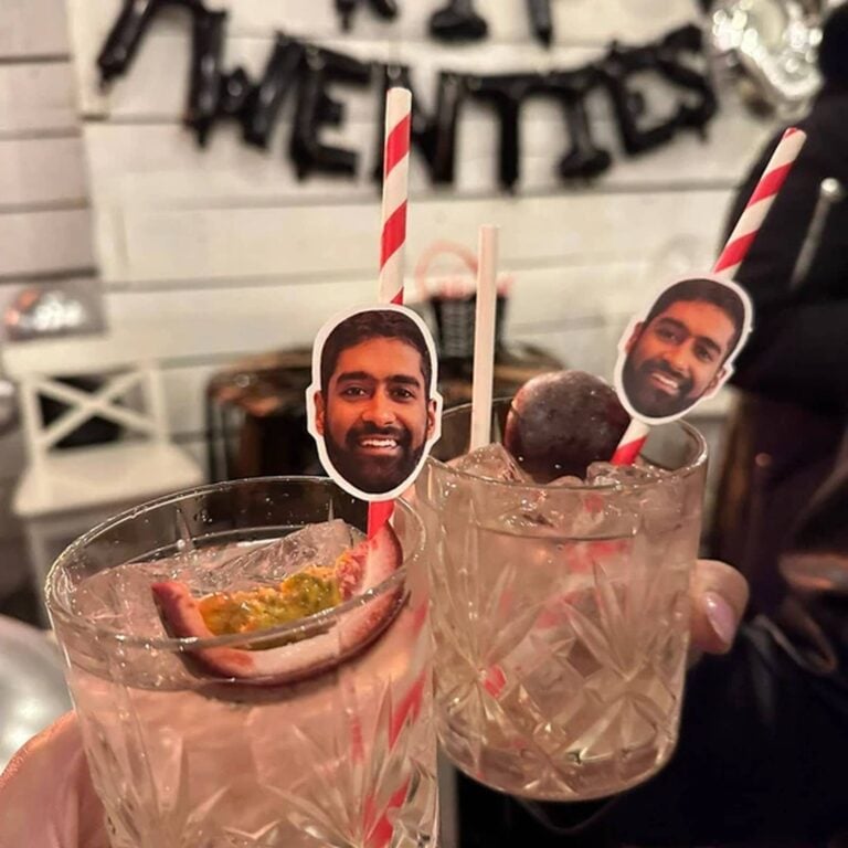 Two cocktails with red-and-white striped photo straws topped with the same man’s smiling face, “Twenties” balloon banner in the background
