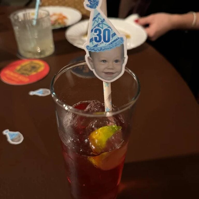 Photo straw with a baby’s face wearing a blue “30” party hat, standing in a dark red cocktail with lime