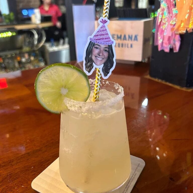 Gold striped photo straw with a smiling woman’s face in a purple party hat, in a salt-rimmed margarita with a lime wheel