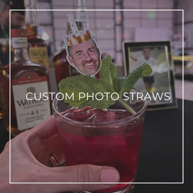 Custom photo straw with a man’s crowned face topper in a red cocktail garnished with mint, whiskey bottles behind, labeled “Custom Photo Straws”
