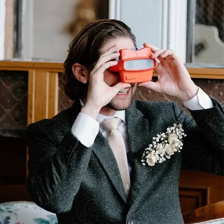 Groom holding an orange reel viewer to his eyes at a wedding, boutonniere on lapel