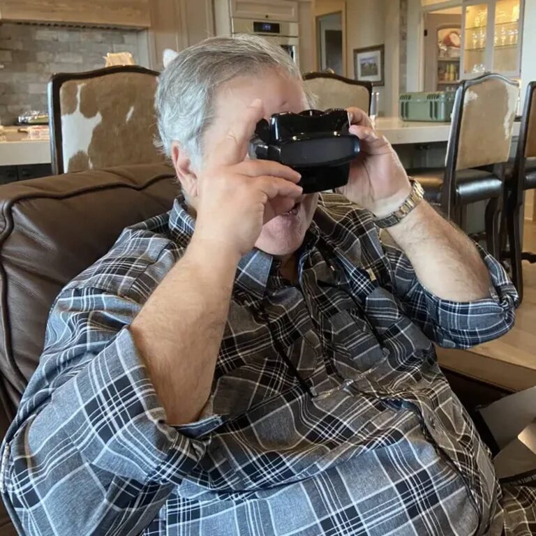 Older man in a plaid shirt looking through a black reel viewer in a kitchen