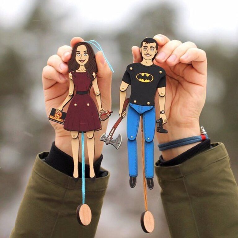Two custom wooden portrait ornaments of a couple — a woman in burgundy dress and a man in Batman t-shirt — held up against a snowy backdrop