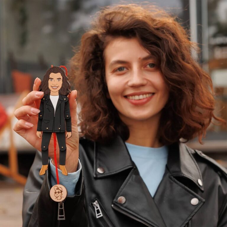 Smiling woman in leather jacket holding a custom wooden portrait ornament of herself in an all-black outfit with curly hair