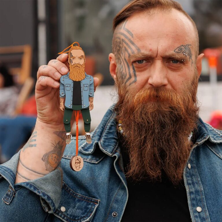 Tattooed man with long red beard holding a custom wooden portrait ornament of himself in a denim jacket and dark trousers