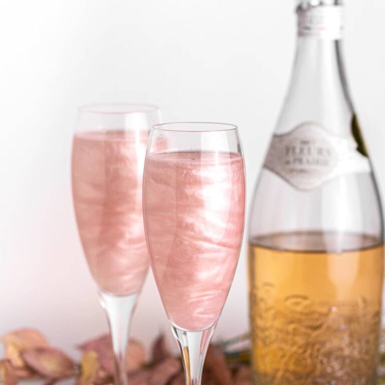 Two champagne flutes of pink edible glitter drink beside a bottle of sparkling wine