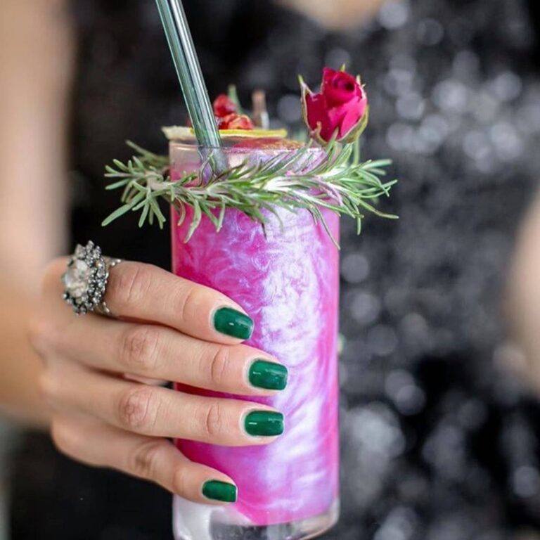 Hand holding a pink edible glitter cocktail garnished with a rosemary sprig and a rose bud