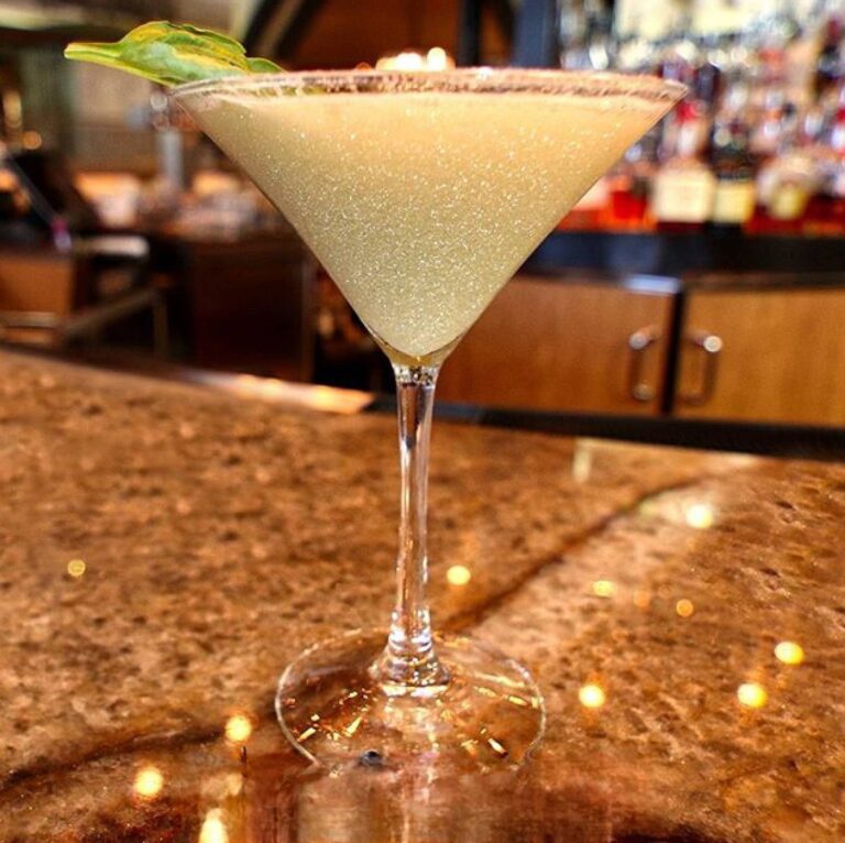 Martini glass with pale gold edible glitter cocktail, a sugared rim, and a lime leaf garnish at a bar