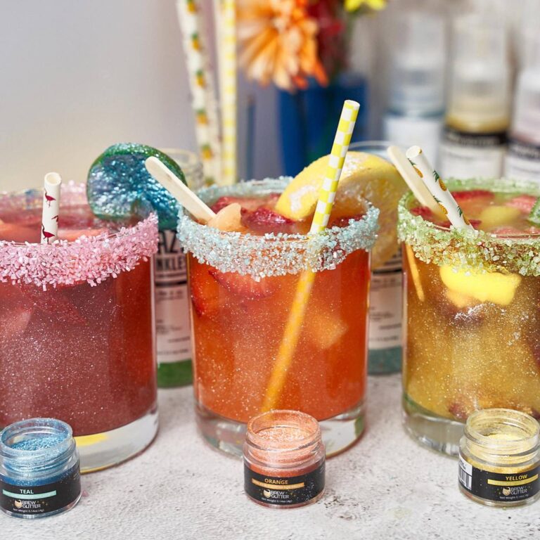 Three edible glitter cocktails with colored sugar rims in teal, orange, and yellow, each beside its matching Brew Glitter jar