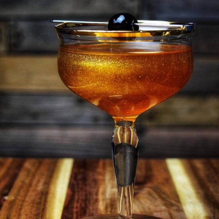 Gold edible glitter cocktail in a coupe glass garnished with a black olive