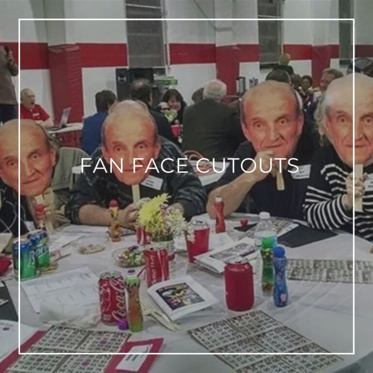 Group of people sat at a table, holding up fan face cutouts of a man in front of their face.