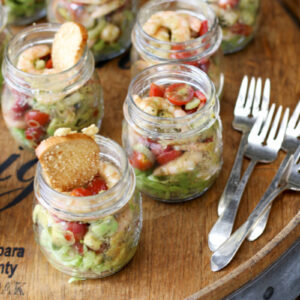 Shrimp and avocado salad mason jars topped with a crostini garnish and silver forks on a wine barrel