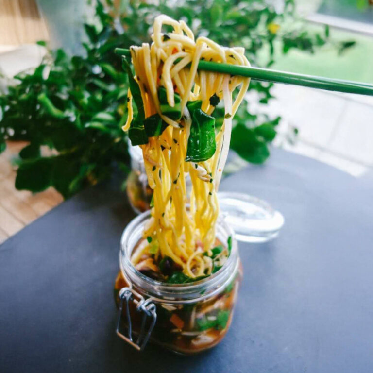 Asian noodle salad mason jar with chopsticks lifting noodles above the jar, styled with greenery in the background