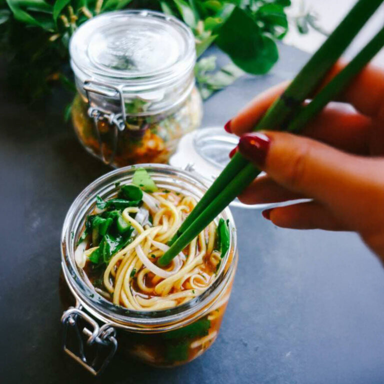 Hand using chopsticks to stir a noodle salad mason jar, with a second jar and greenery in the background